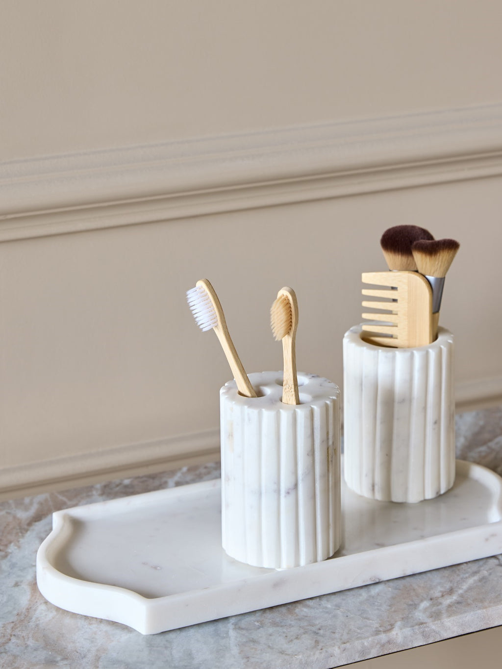 Fluted white marble holder containing two wooden toothbrushes on a pale tray beside a matching vessel holding brushes and a comb.