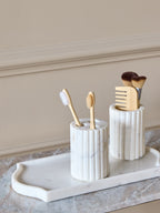 Fluted white marble holder containing two wooden toothbrushes on a pale tray beside a matching vessel holding brushes and a comb.