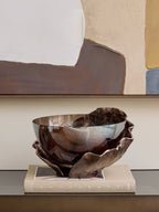 Smoked glass bowl with a folded bronze-toned surround displayed on a beige book beneath a large abstract artwork.