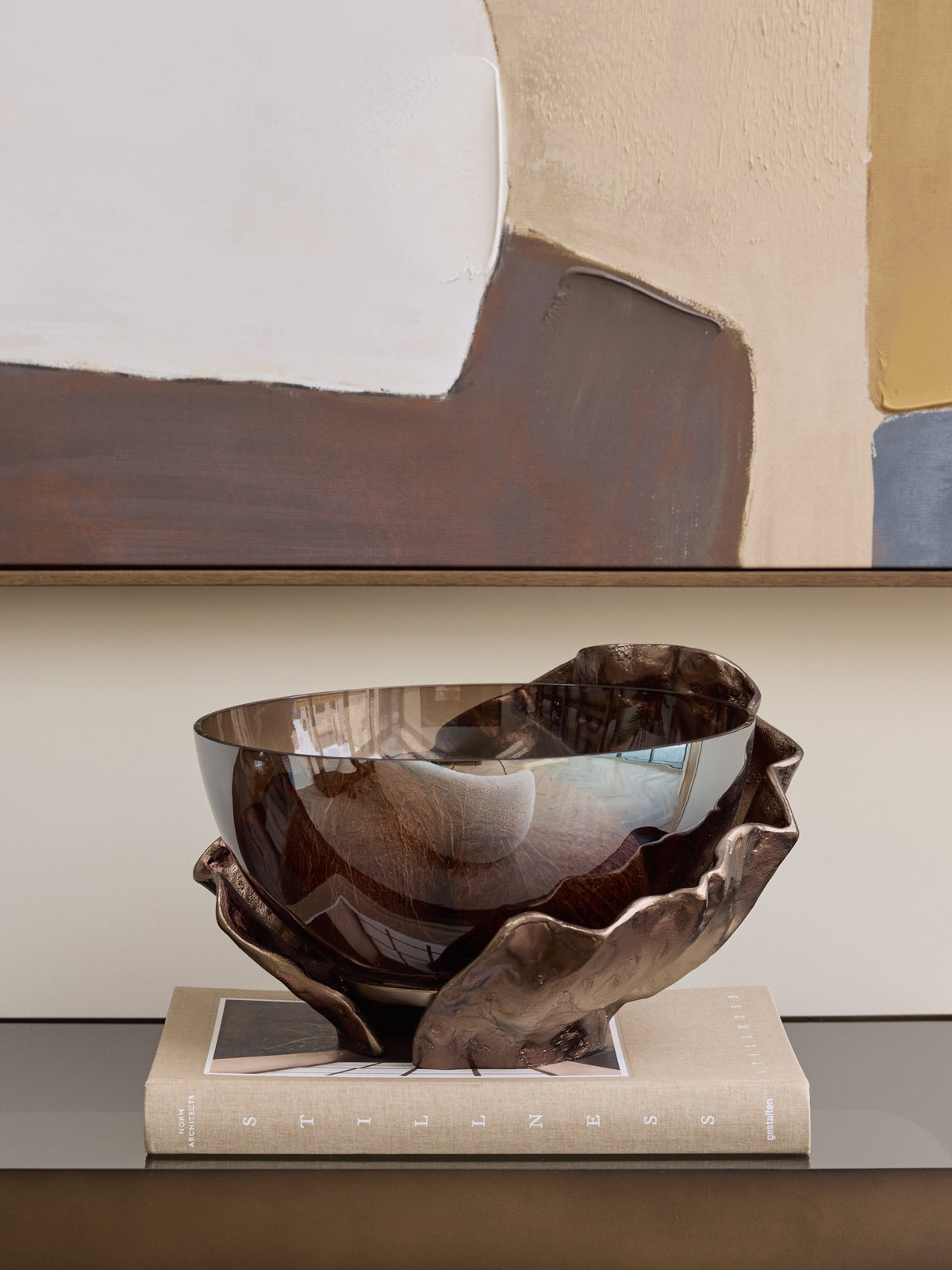 Smoked glass bowl with a folded bronze-toned surround displayed on a beige book beneath a large abstract artwork.