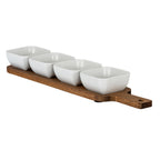 Angled view of four glossy white rounded-square bowls arranged on a warm brown paddle-shaped board, with its handle extending to the right.