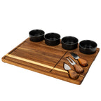 Angled view of a brown grained cheese board with four dark bowls along the back and three rounded-handle utensils in slots on the right.