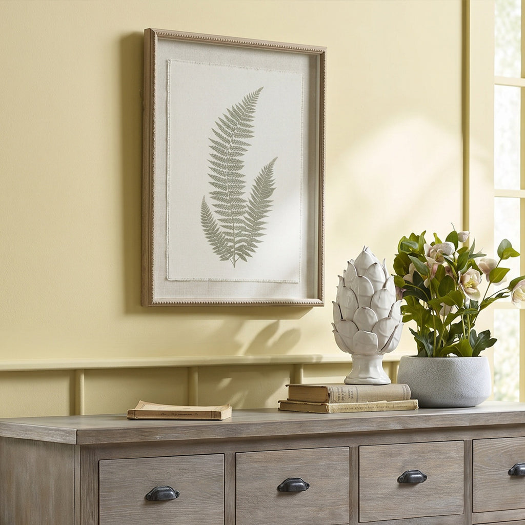 Three-frond botanical artwork hanging on a pale yellow wall above a grey-brown sideboard, beside books, a white ornament and flowering foliage.