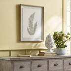 Three-frond botanical artwork hanging on a pale yellow wall above a grey-brown sideboard, beside books, a white ornament and flowering foliage.