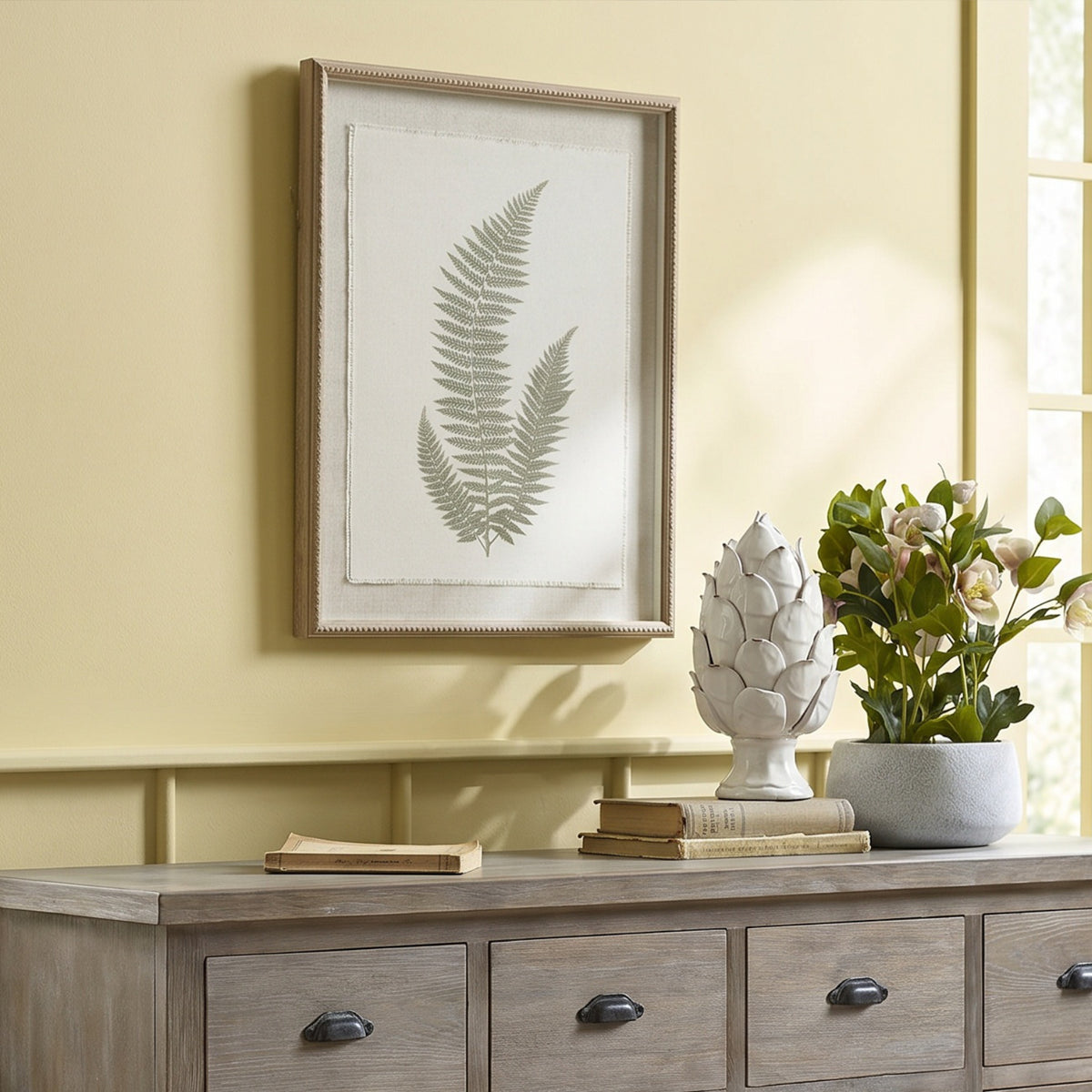 Three-frond botanical artwork hanging on a pale yellow wall above a grey-brown sideboard, beside books, a white ornament and flowering foliage.