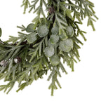 Close-up of frosted rounded leaves and fine green sprigs, with brown pinecones and part of the twig-like wreath ring visible.