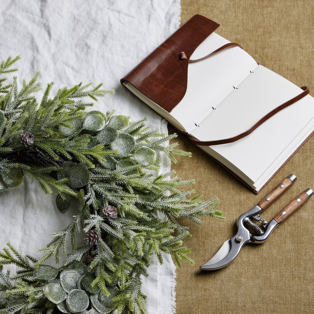 Frosted foliage wreath resting on white and tan fabric beside an open brown-covered notebook and pruning shears.