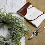 Frosted foliage wreath resting on white and tan fabric beside an open brown-covered notebook and pruning shears.