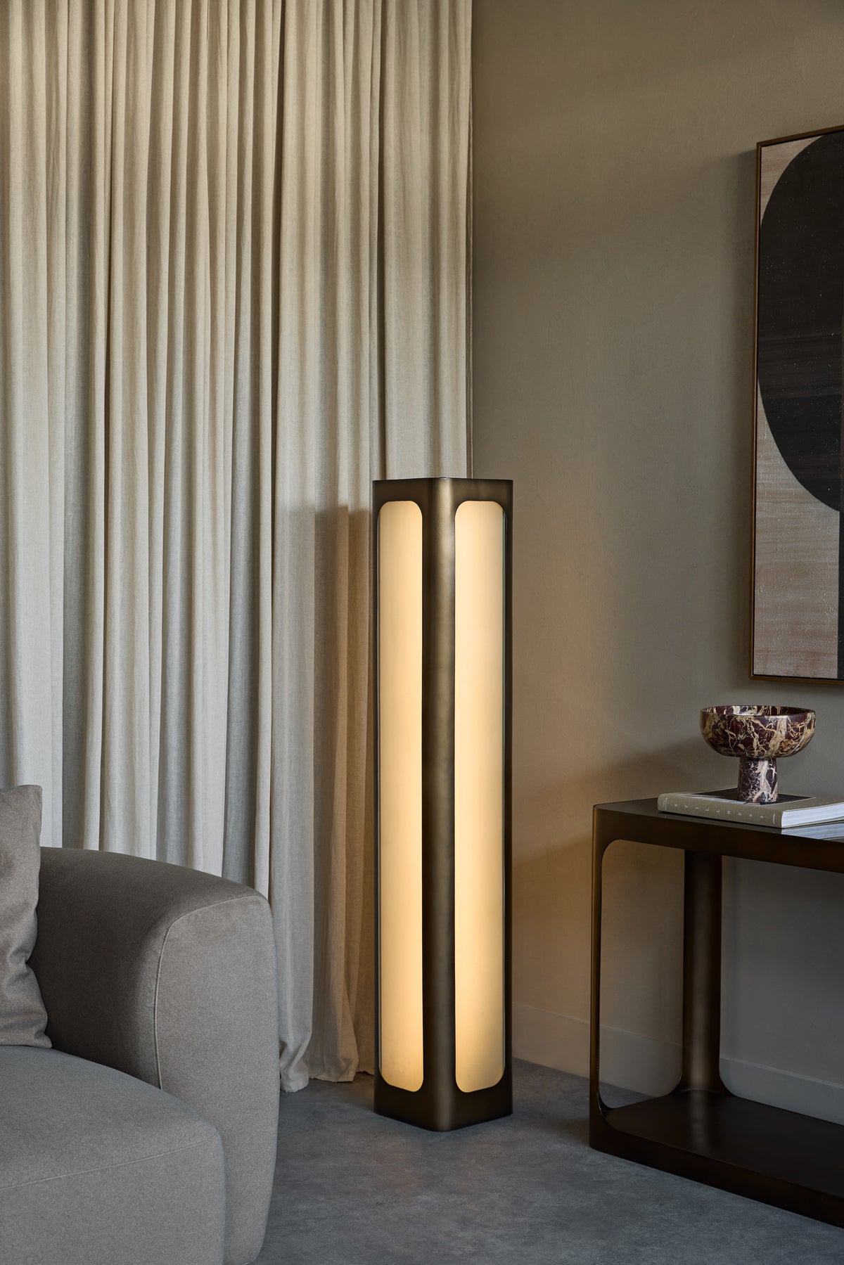 Illuminated frosted-panel floor uplighter beside cream curtains, a grey sofa and a dark console table in a neutral living room.