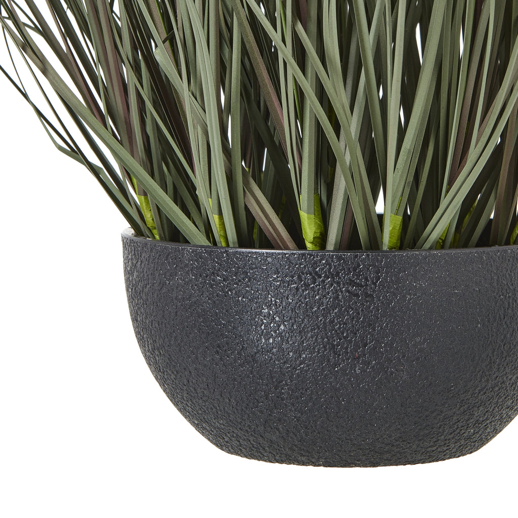 Close-up of the pebbled black bowl-shaped pot beneath the lower green blades of the artificial grass arrangement.