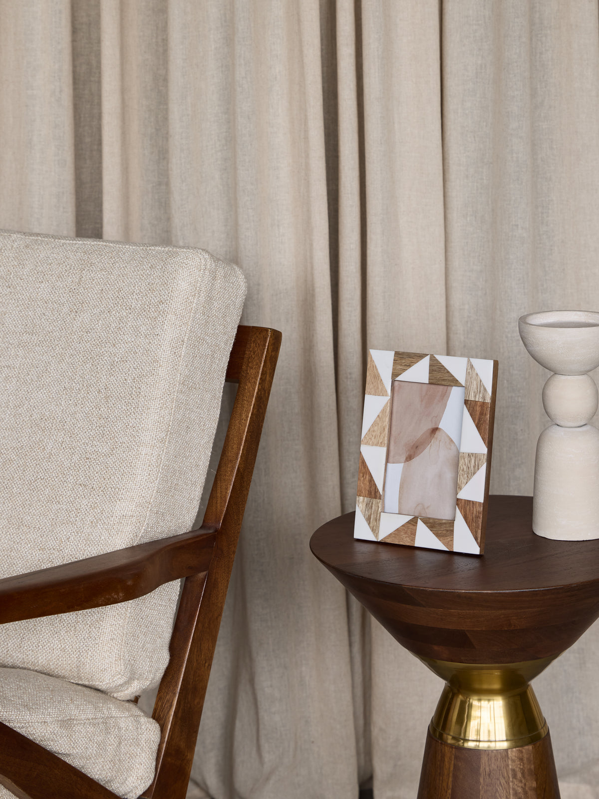 Brown and cream triangular-patterned photo frame on a dark wooden side table beside a pale sculptural ornament and an upholstered wooden armchair.