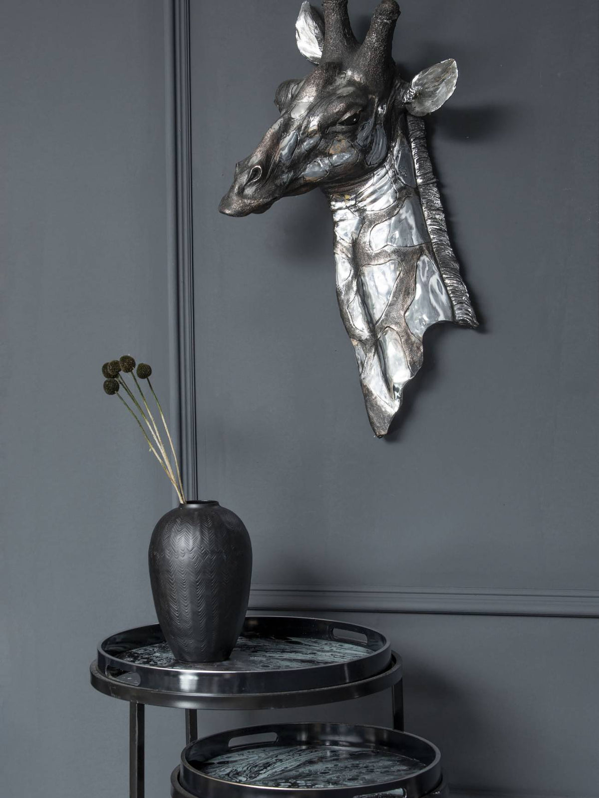 Black and silver giraffe plaque displayed on a dark grey panelled wall above round tray tables and a black vase with stems.