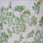 Detail of pale green flowers and curling leaves with darker green accents, fine crackling and tan marks on a cream-white background.