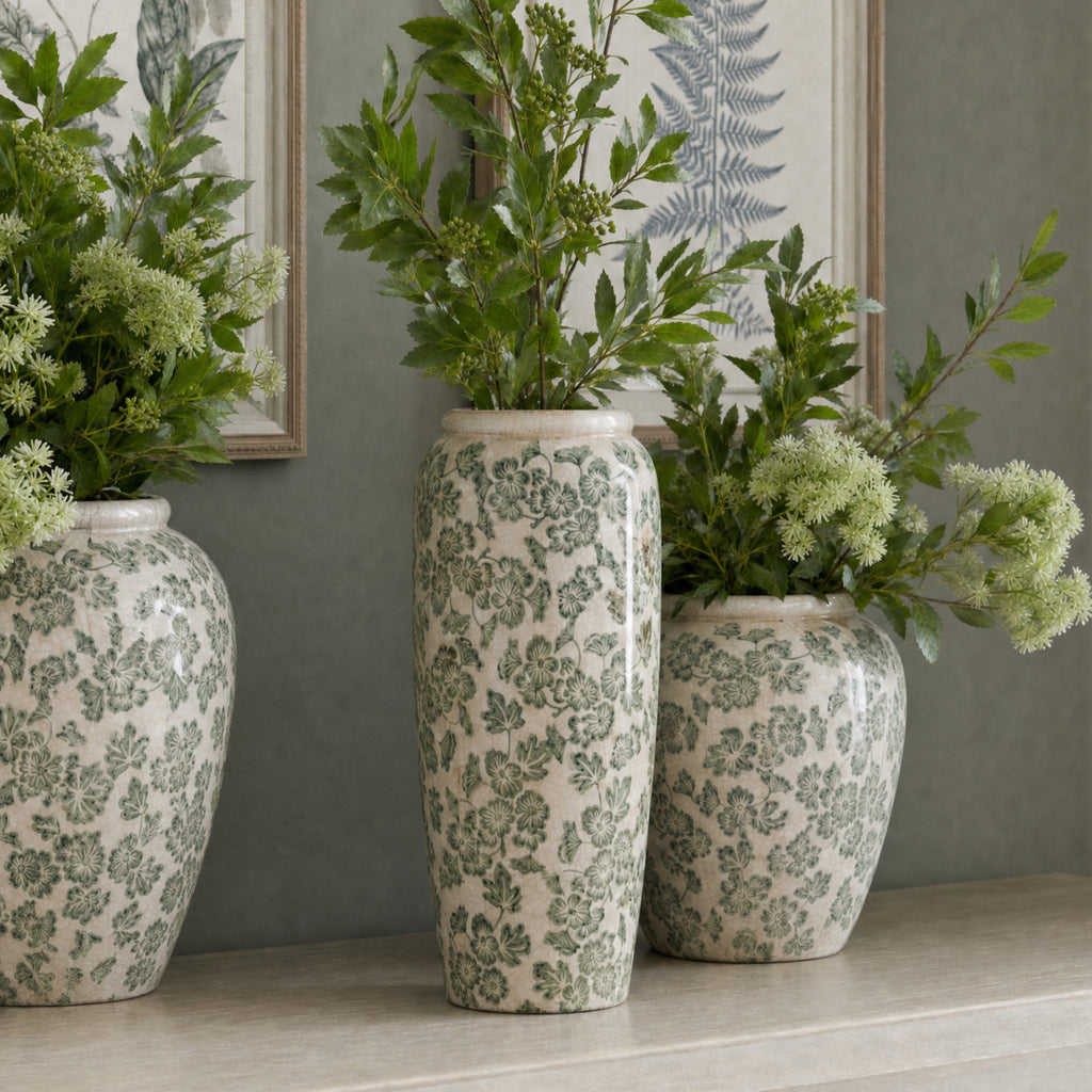Tall green floral vase filled with leafy stems on a console, between two rounder patterned vases beneath framed botanical prints.