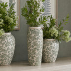 Tall green floral vase filled with leafy stems on a console, between two rounder patterned vases beneath framed botanical prints.