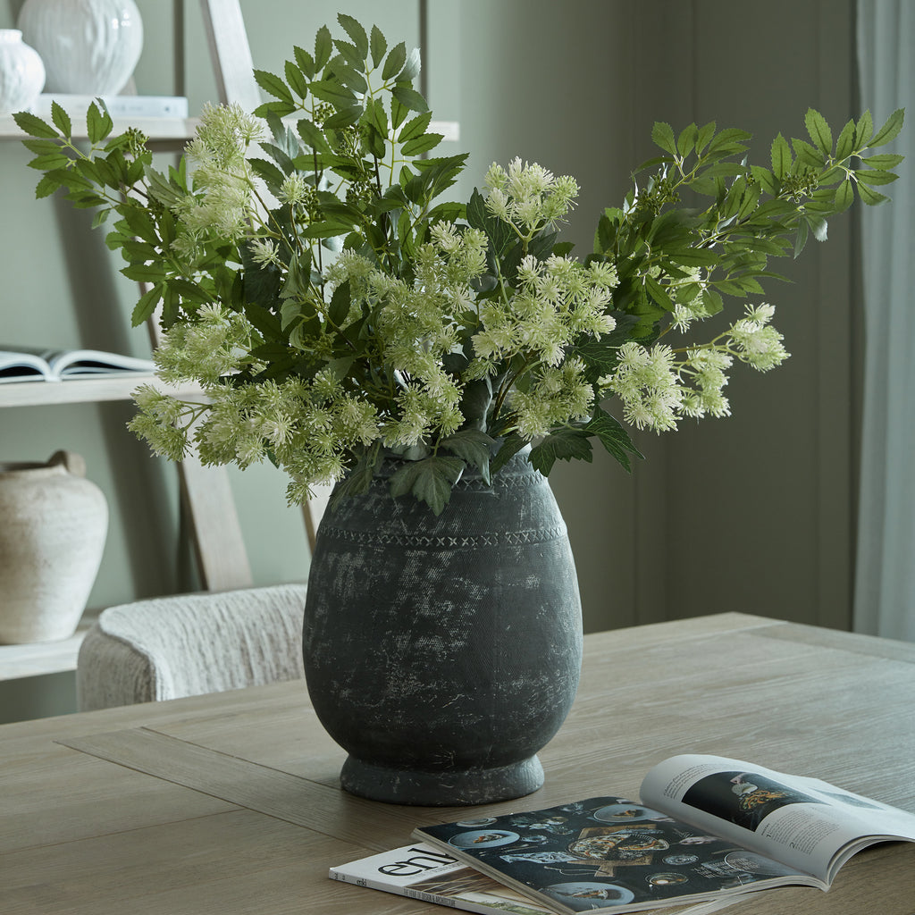 Styled arrangement of pale meadow flower clusters and additional green foliage in a dark rounded vase on a wooden table beside open magazines.