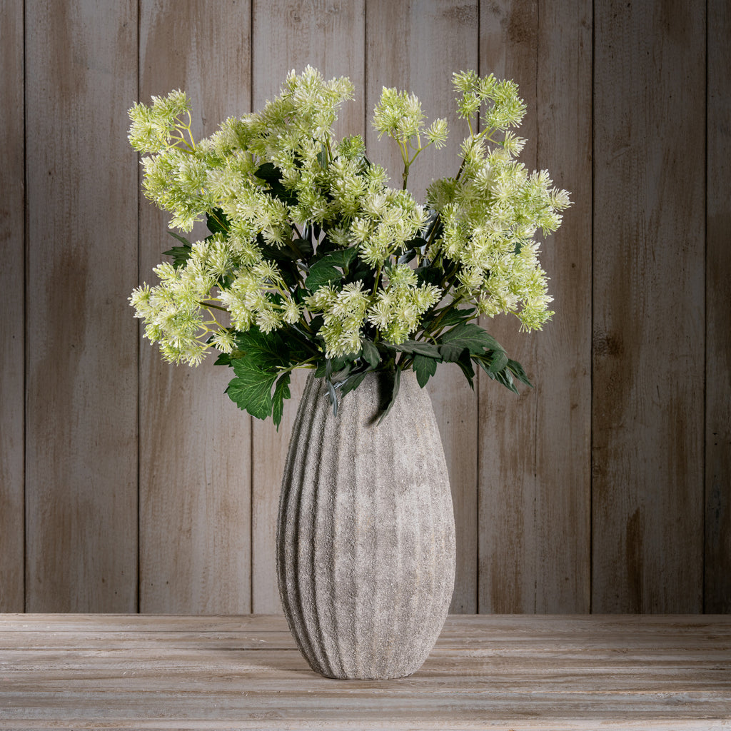 Styled arrangement of pale green meadow flower sprays in a tall textured grey vase on a wooden surface against vertical wood panelling.