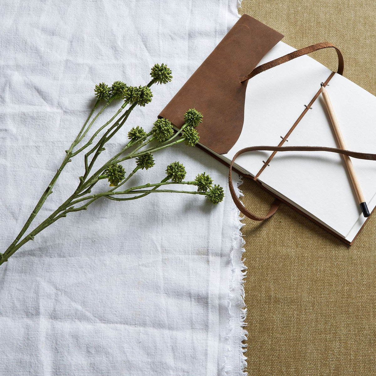 Green artificial flower stem laid diagonally on fringed white cloth beside an open notebook and a wooden-coloured pen.