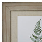 Close-up of the artwork's upper-left frame corner, showing its wood-grain appearance, beige woven-looking surround and the tip of a green fern frond.