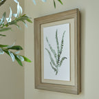 Angled view of the framed green botanical artwork hanging on a pale wall, with leafy branches in the foreground.