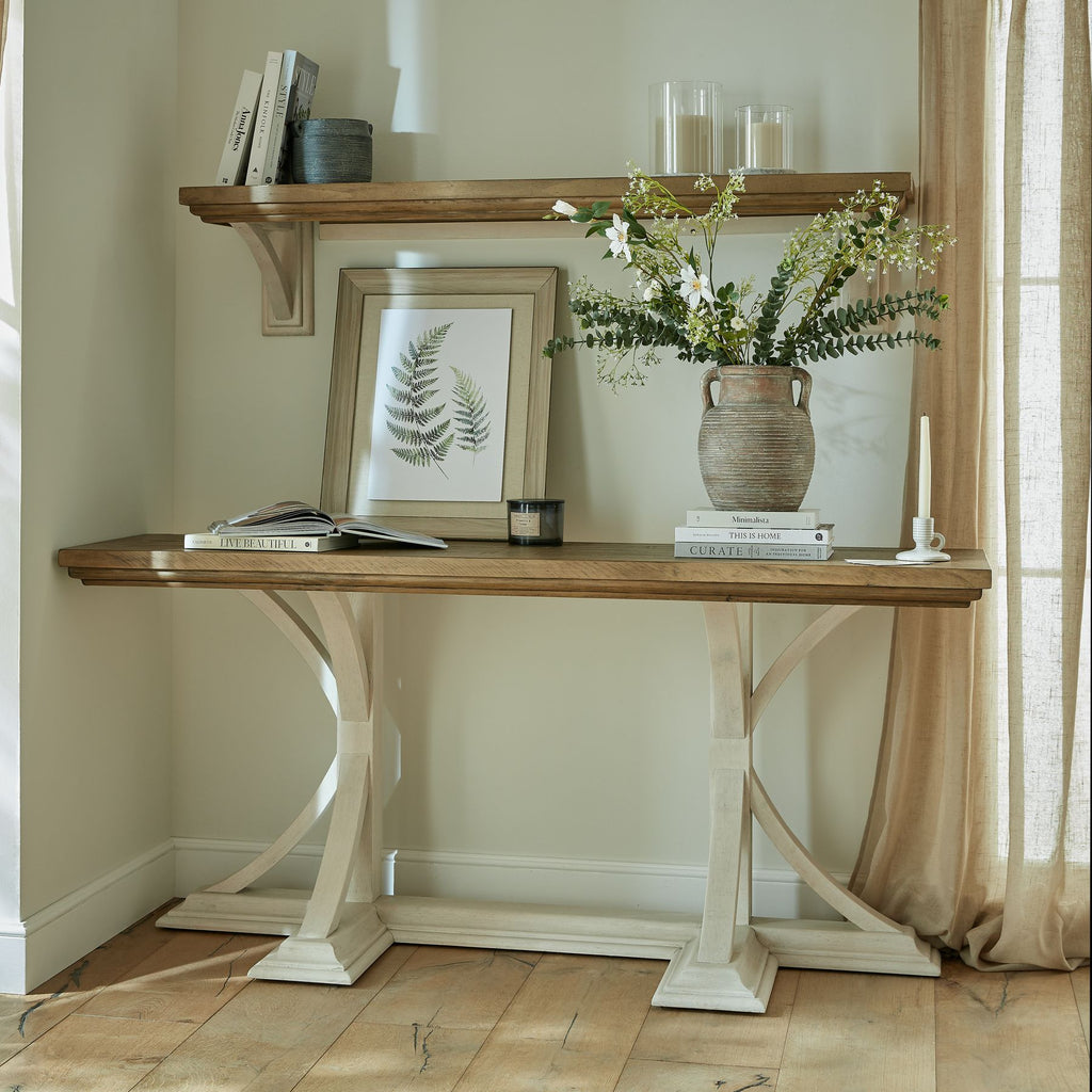 Two Fronds artwork leaning on a console beneath a wall shelf, beside books, candles and a handled vase of green stems and white flowers.