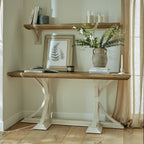 Two Fronds artwork leaning on a console beneath a wall shelf, beside books, candles and a handled vase of green stems and white flowers.