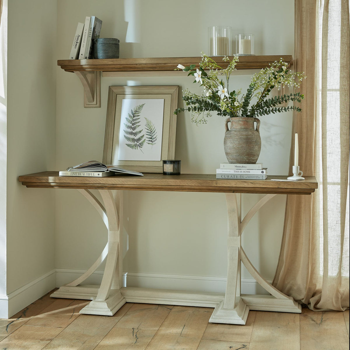 Two Fronds artwork leaning on a console beneath a wall shelf, beside books, candles and a handled vase of green stems and white flowers.