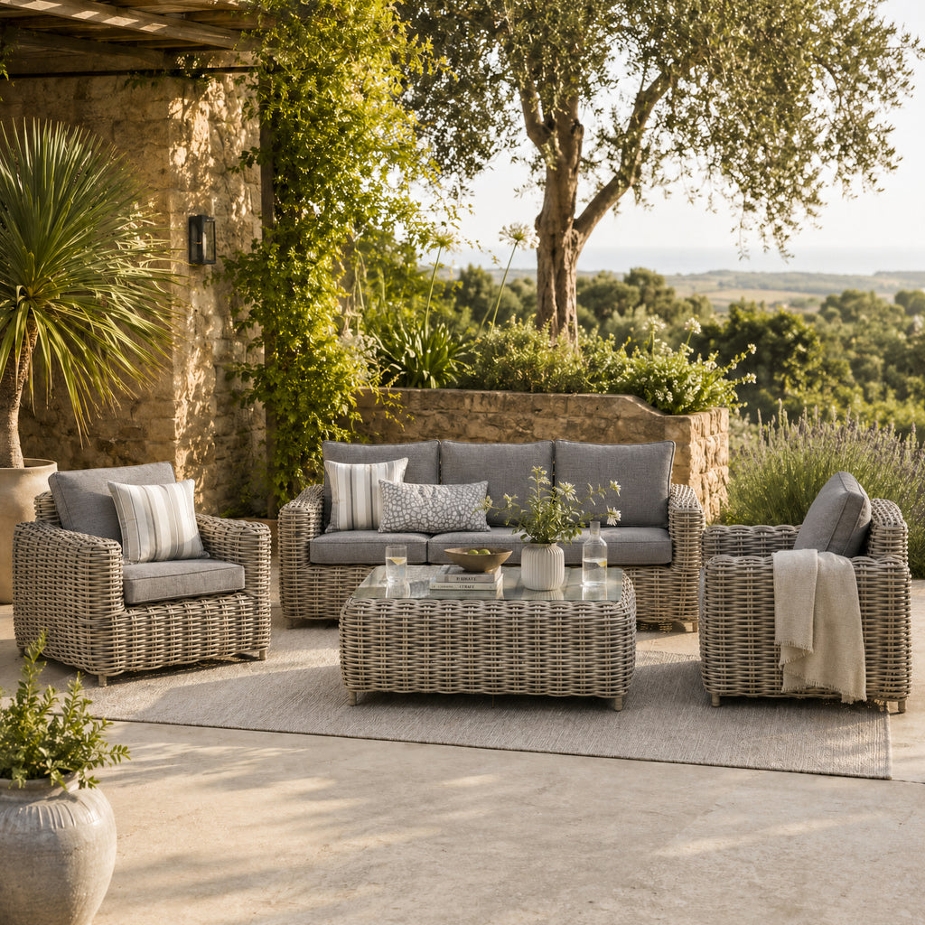Grey woven sofa and two armchairs arranged around a low rectangular table on a cream rug, styled with cushions and flowers on a stone terrace.