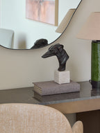Greyhound sculpture displayed on a fabric-covered book beneath a round mirror, beside a green lamp on a console.