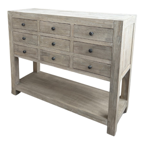 Three-quarter view of a pale grey-brown chest with nine drawers, dark drawer hardware, broad corner posts and an open lower shelf against a white background.