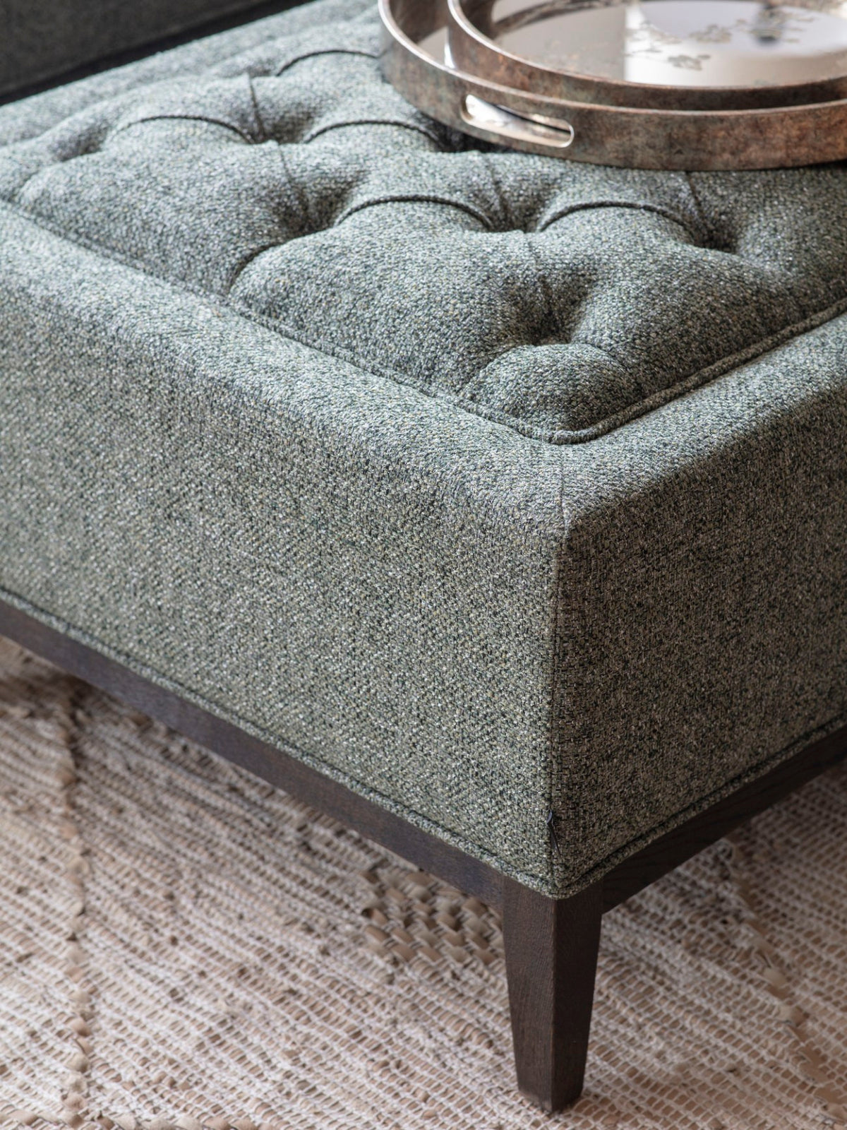 Close-up of the ottoman's textured green weave, recessed buttons, piped corner and tapered wooden leg, with round trays on top.