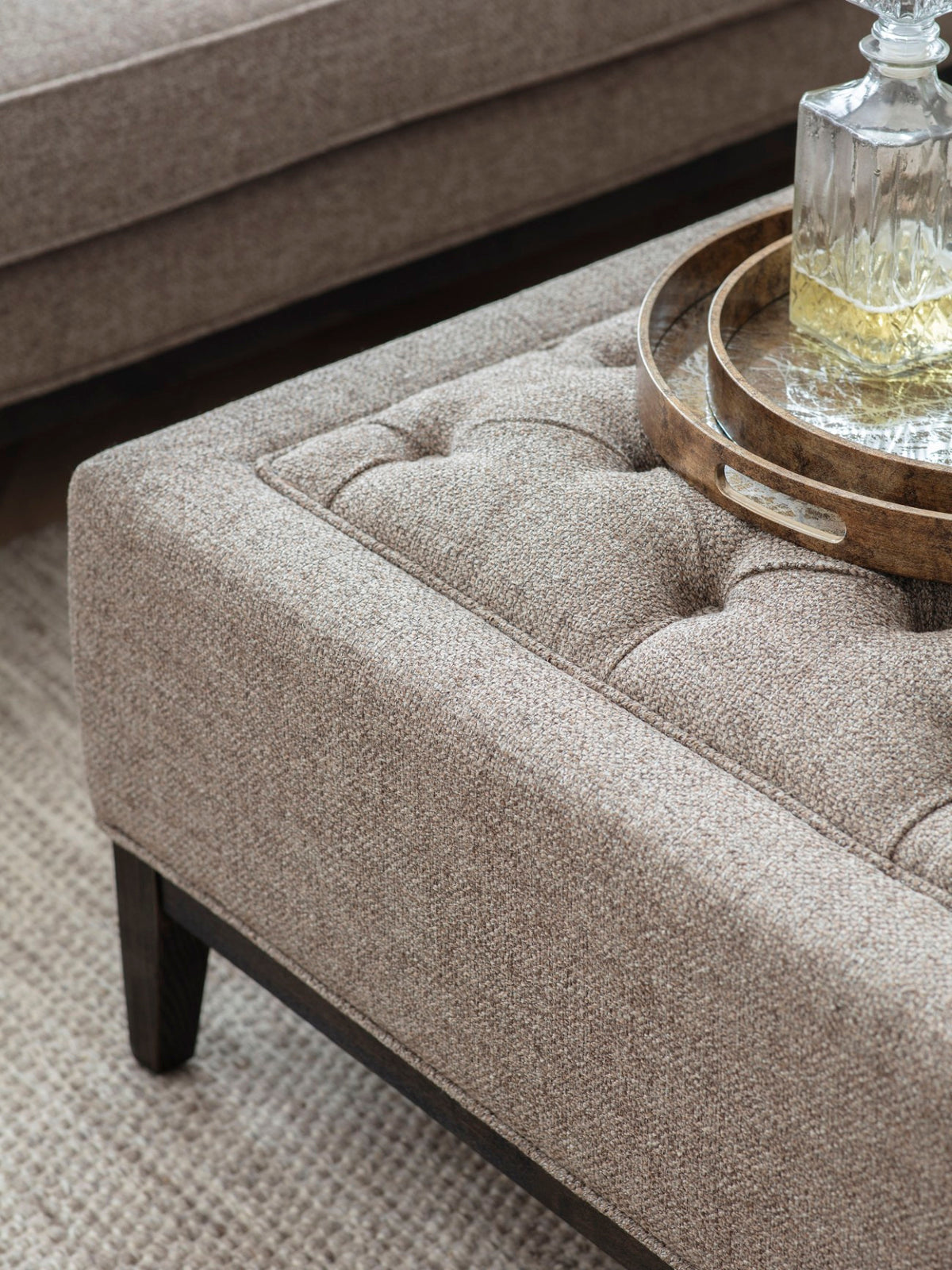 Close-up of the ottoman’s textured taupe weave, deep buttoning and piped border, with round trays and a glass decanter on top.