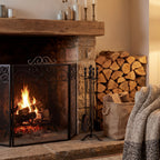 Heart and scroll fire screen pictured before a lit stone fireplace beneath a wooden mantel, beside fireplace tools, stacked logs and a knitted throw.