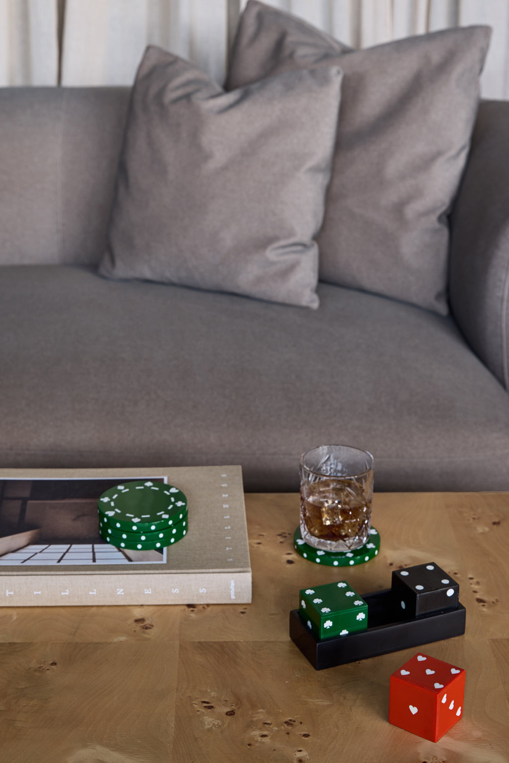 Dice set on a wooden coffee table in front of a grey sofa, with the red heart die outside the tray beside books, green coasters and a drinking glass.