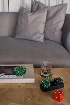Dice set on a wooden coffee table in front of a grey sofa, with the red heart die outside the tray beside books, green coasters and a drinking glass.