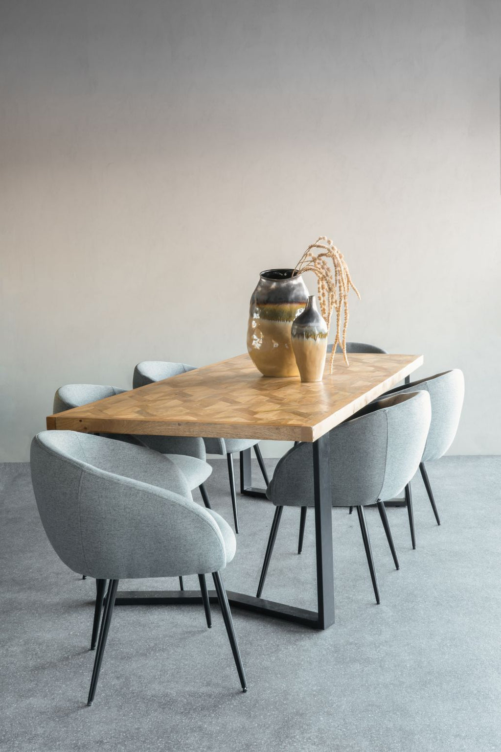 Herringbone dining table surrounded by grey upholstered chairs, styled with two ceramic vases and dried stems.