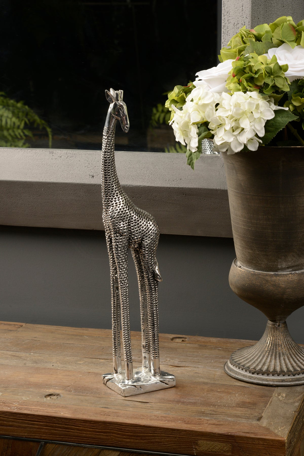 Silver giraffe sculpture on a wooden console beside a large urn of white and green flowers, with a mirror behind.
