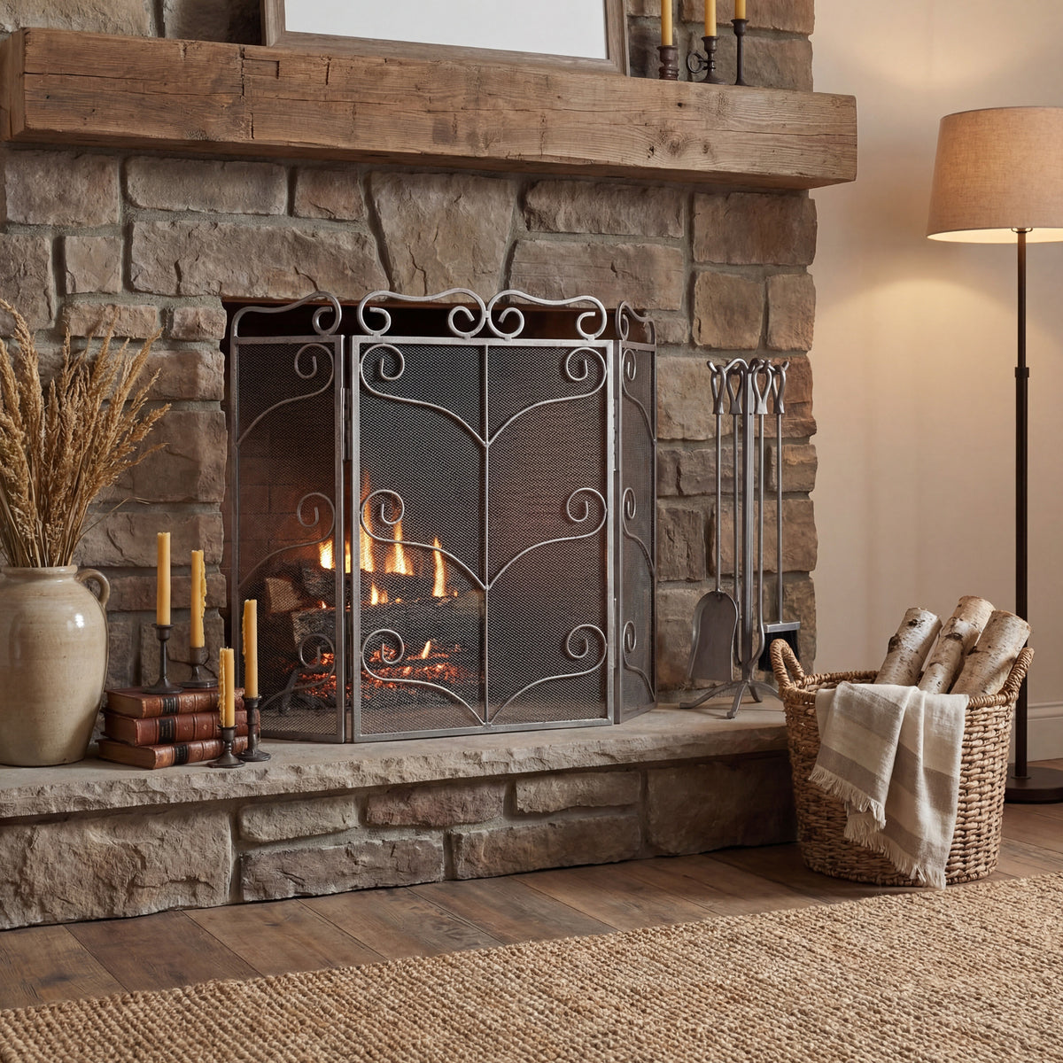 Scroll fire screen in front of a lit stone fireplace beneath a timber mantel, styled with candles, a ceramic vase, fireside tools and a basket of logs.