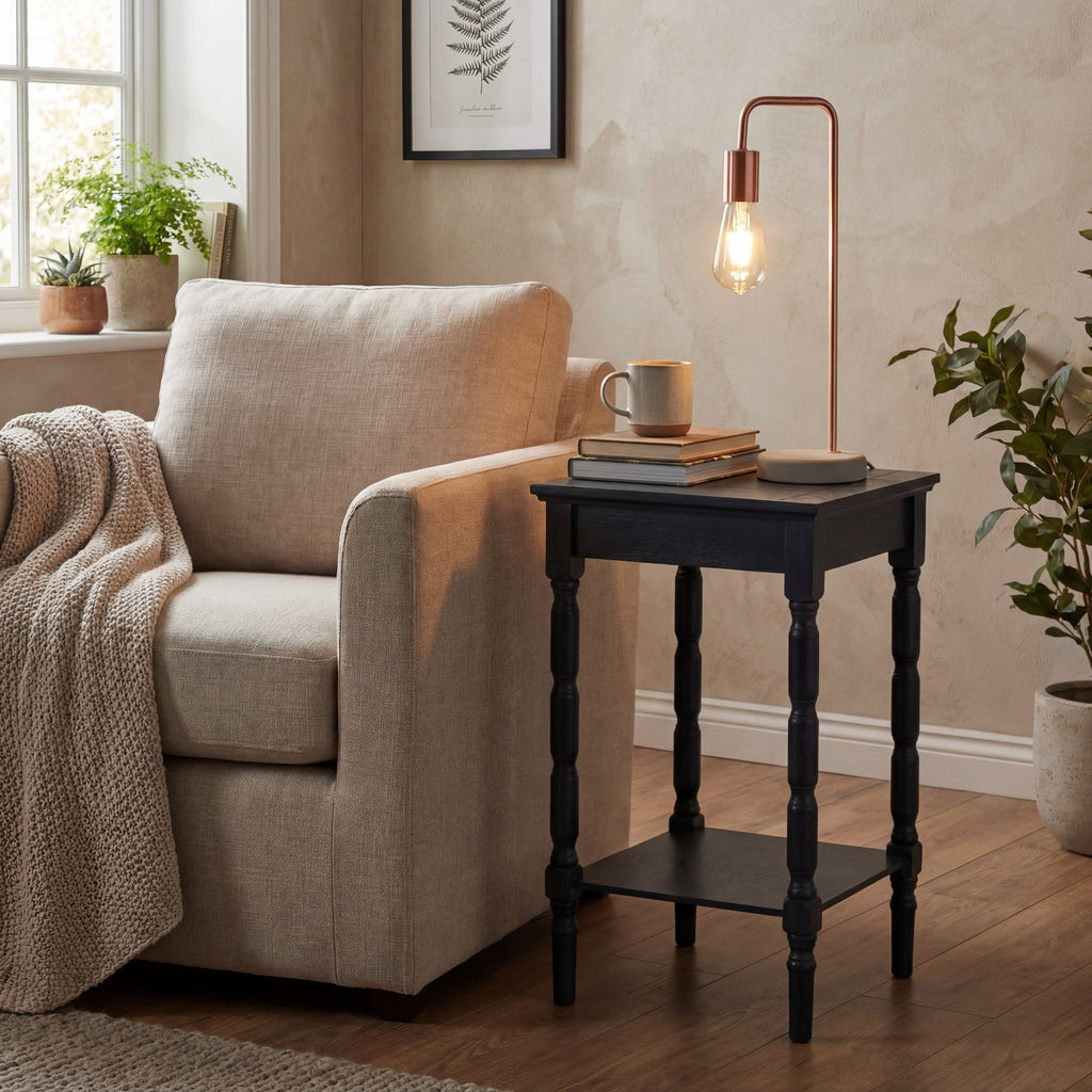 Holder Frame Table Lamp with a glowing bulb on a dark side table beside a cream armchair, styled with books and a mug in a neutral room.