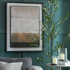 Horizontal Fields Framed Artwork displayed on a deep green panelled wall above a grey sofa, beside leafy branches and candles on a round side table.