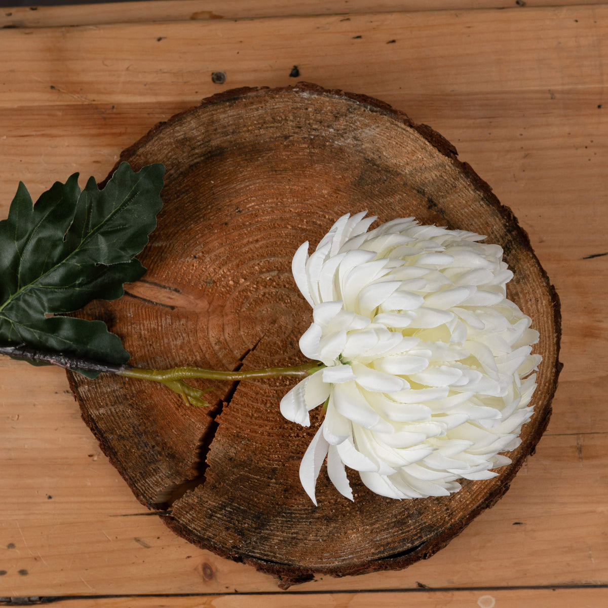 Close-up of layered off-white chrysanthemum petals and dark green leaves, with the artificial stem laid across a wooden tree slice.