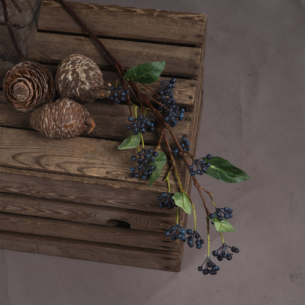 Indigo berry spray laid diagonally across a wooden slatted surface beside three pine cones, with leafy branches extending over the edge.