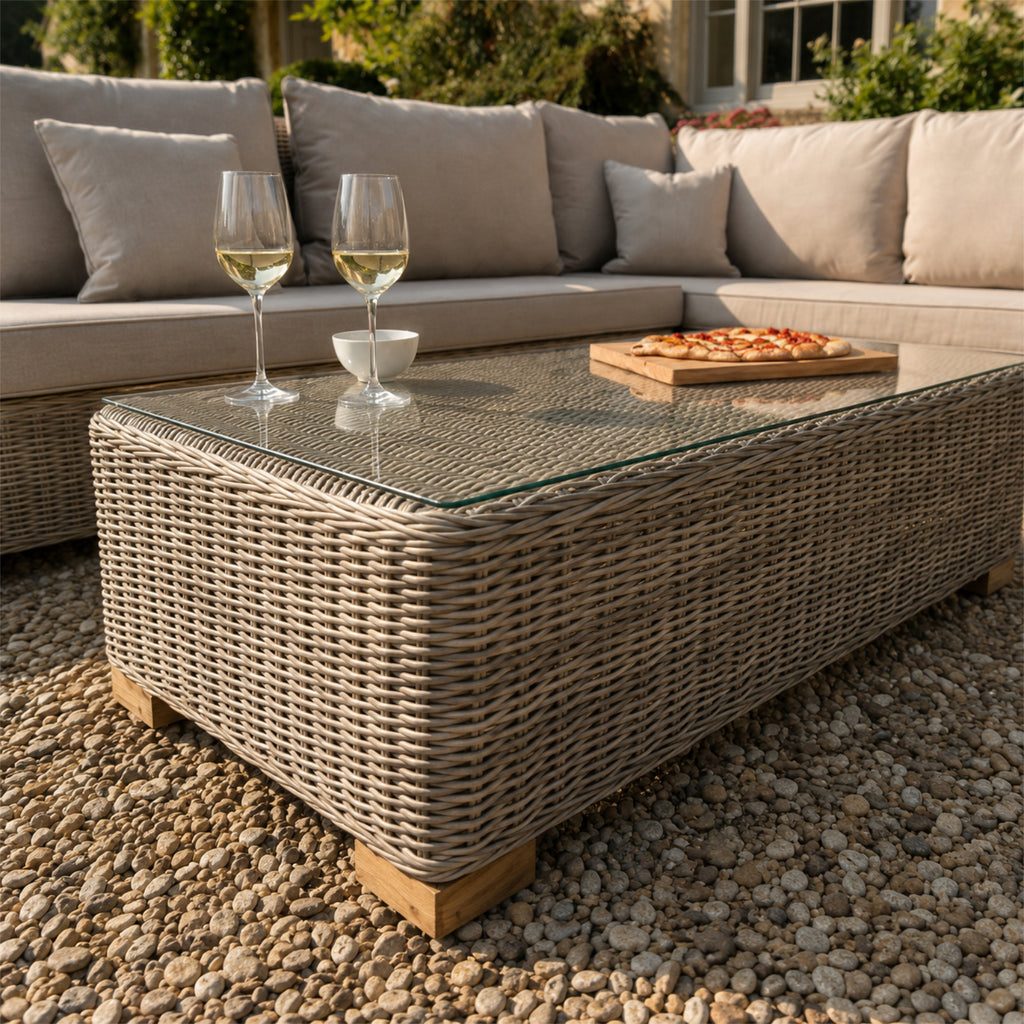 Close view of the woven coffee table with a transparent-looking top and small block feet, styled with wine glasses, a bowl and pizza in front of pale cushioned seating.