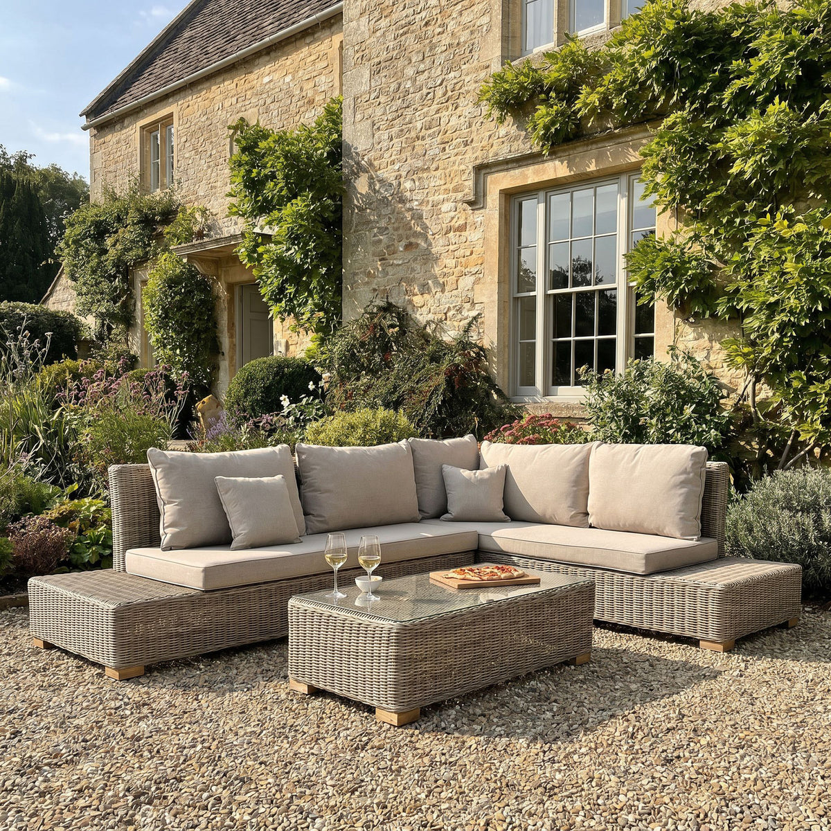 Woven corner seating and coffee table photographed on gravel beside a stone house and leafy planting, with pale cushions and food and drinks arranged on the table.