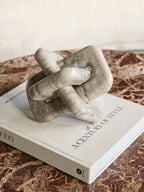 Beige marble knot sculpture resting on a cream art book on a richly veined reddish-brown stone table.