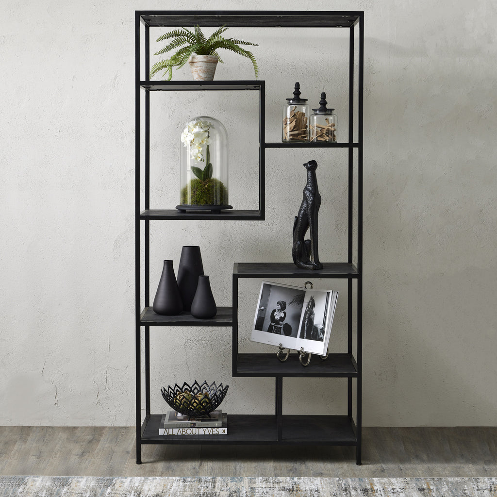 Black shelving unit against a pale textured wall, styled with a fern, glass jars, flowers under a glass dome, black vases, an animal sculpture and an open book.