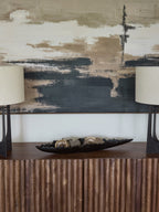 Black irregular-edge dish holding neutral decorative objects on a fluted wooden sideboard between two lamps beneath abstract artwork.