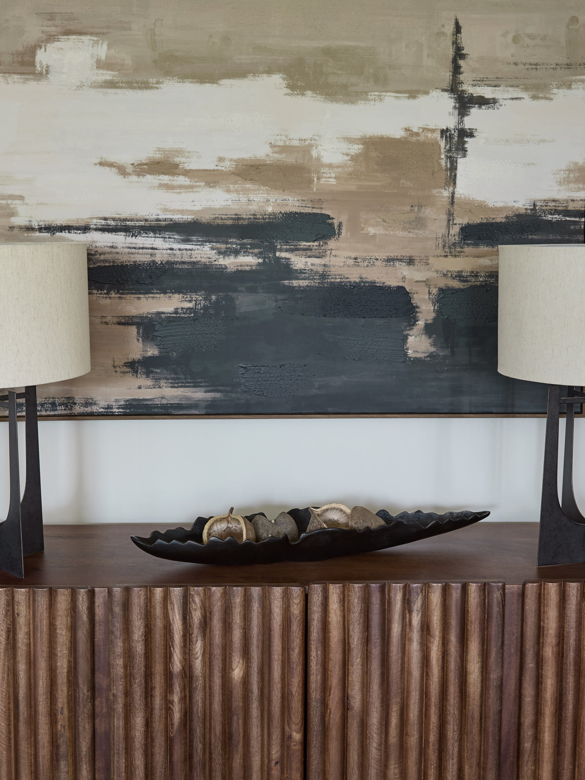 Black irregular-edge dish holding neutral decorative objects on a fluted wooden sideboard between two lamps beneath abstract artwork.