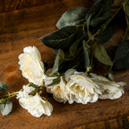 Ivory artificial flowers lying diagonally on a brown wooden surface, with overlapping petals, small buds and dark green veined foliage.