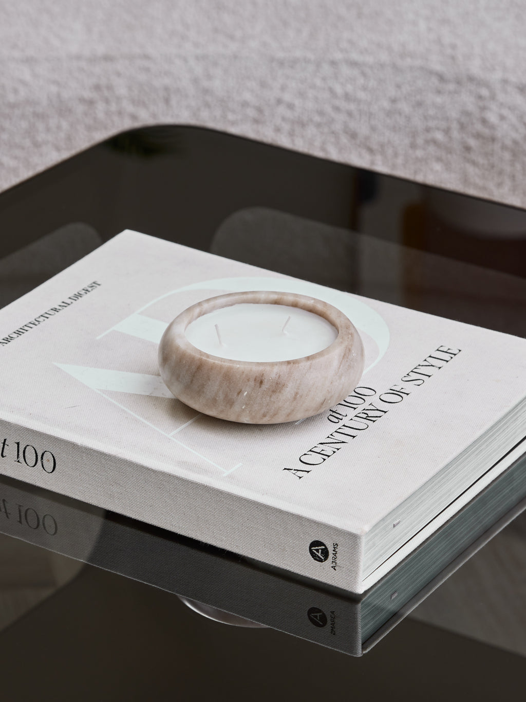 Beige marble candle with two unlit wicks resting on an Architectural Digest book on a reflective dark glass table.